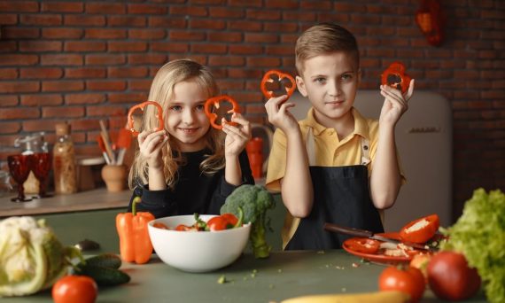Two children holding capsicum slices while preparing vegetables in a cozy kitchen setting.